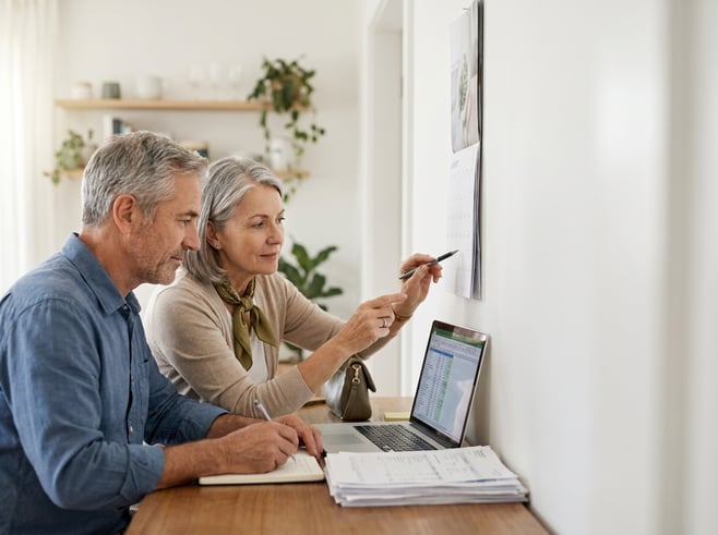 A thoughtful older couple seated at a dining table reviewing financial papers and a calendar with a laptop nearby, lit by soft morning window light in a tidy home setting. The image feels calm and organized, focused on collaboration and year-end planning.