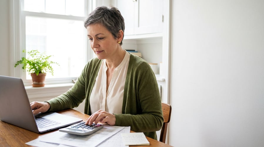 A senior woman in her early 60s sits at a bright kitchen table reviewing retirement paperwork with a laptop, notes, and a calculator nearby, lit by soft window light for a calm, focused planning scene.